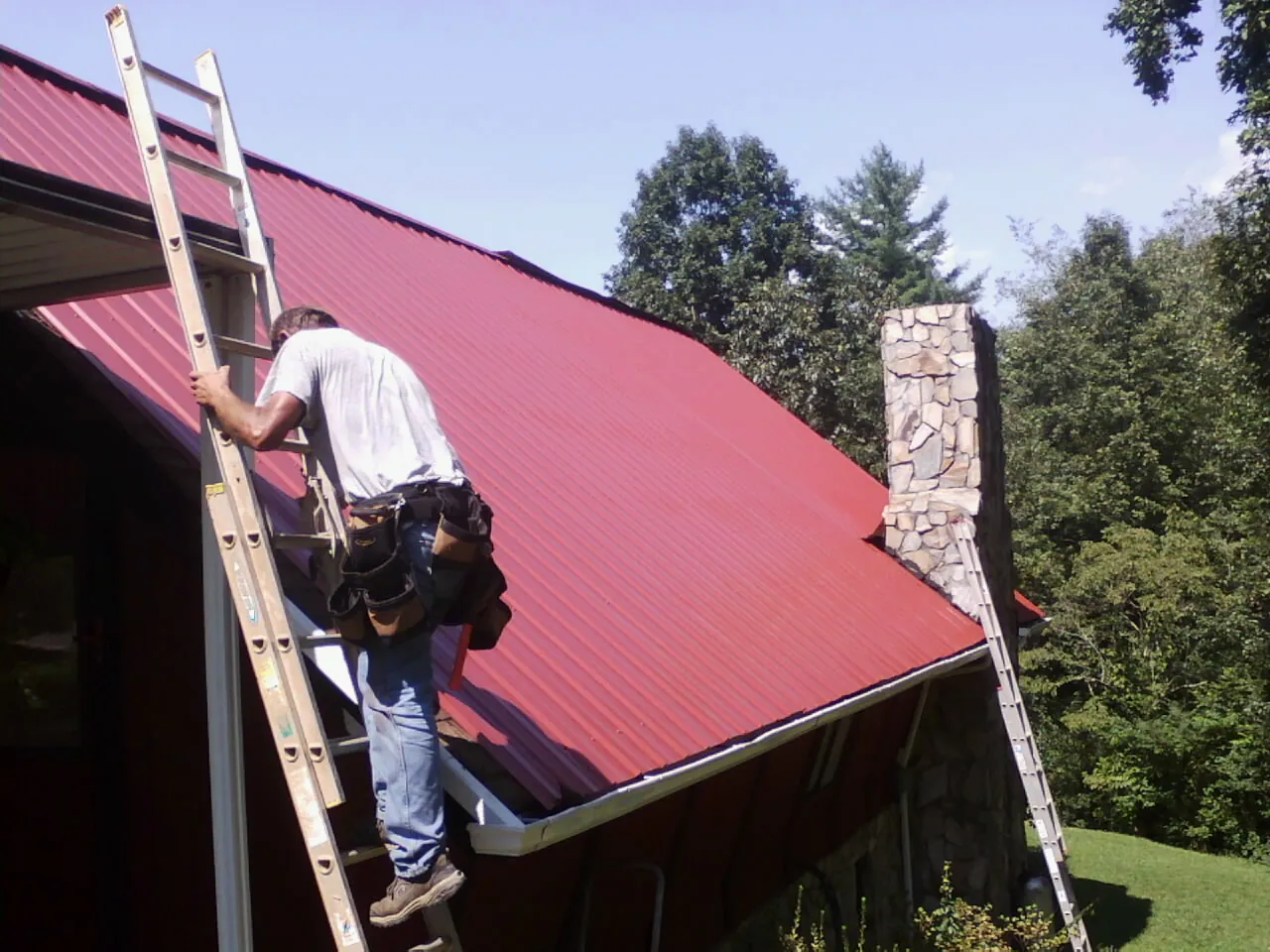 Roofer on a ladder beside a red metal roof and stone chimney — Soffit & Fascia Repair by Roofing Brothers in Montpelier